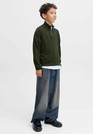 Zeleni zip-up flis pulover, bel majica pod njim, široke izbledele modre jeans hlače in črne čevlje. Ima visok ovratnik in sproščen kroj.