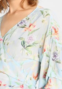 Květinový top s obálkovým střihém vyrobený z světle modré látky. Má živé, vícebarevné květinové vzory, splývavé rukávy a výstřih do V.