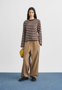 Nő áll kék szőnyegen, barna csíkos hosszú ujjú inget, bézs bő szárú nadrágot és fekete cipőt visel, bal kezében sárga sapkát tart.