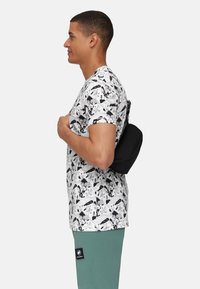 Borsa a tracolla nera su una t-shirt bianca con illustrazioni in stile fumetto nere; il modello indossa pantaloni verdi con una toppa del marchio.