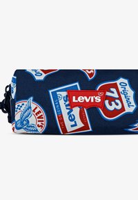 Mėlynas kanvos maišelis su įvairiomis grafinėmis pleistrais, įskaitant raudonus ir baltus Levi's logotipo pleistrus bei sparnų dizainus, turintis užtrauktuko uždarymą.