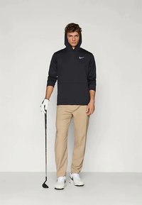 Νεαρός άνδρας που φοράει μαύρη κουκούλα και μπεζ παντελόνι, κρατώντας ένα γκολφ στικάκι, στέκεται μπροστά σε ουδέτερο φωτισμένο φόντο.