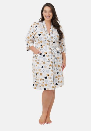 Kobieta uśmiechająca się, ubrana w biały, sięgający kolan szlafrok z czarnymi, beżowymi i szarymi wzorami serc, stojąca boso z rękami w kieszeniach.