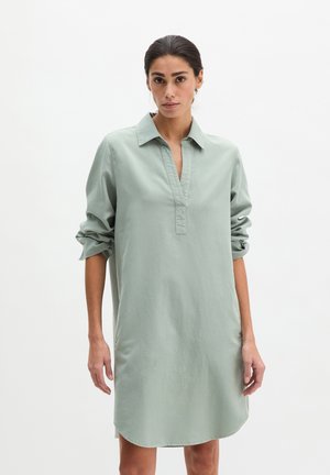 Vrouw met een lichtgroene shirtjurk met lange mouwen, waarvoor de mouwen zijn opgerold, staand tegen een effen witte achtergrond.