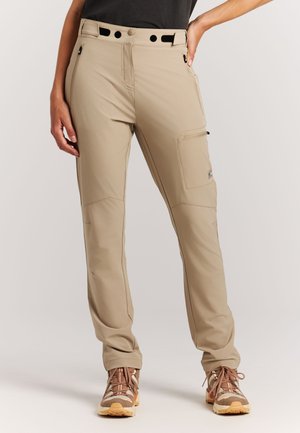 Beige Utility-Hose in schmaler Passform mit Reißverschlusstaschen, getragen mit braunen Wanderschuhen, Beine leicht auseinander und eine Hand auf der Hüfte ruhend.