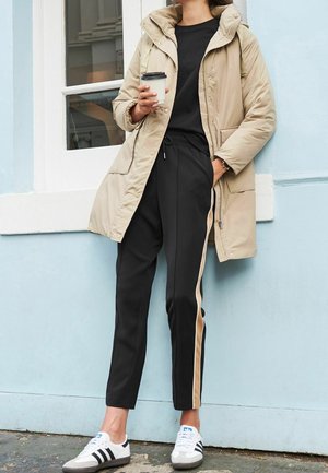 Personne portant un manteau matelassé beige, un haut noir, un pantalon noir avec une bande beige, des baskets blanches Adidas, tenant une tasse de café, appuyée contre un mur bleu pâle.