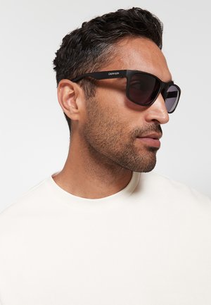 Uomo con capelli corti e scuri e barba curata che indossa occhiali da sole neri Calvin Klein e una maglietta bianca a girocollo, volto verso destra.