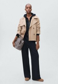 Tan double-breasted trench coat over a black zip hoodie, paired with dark wide-leg pants and light brown slip-on shoes. Gray shoulder bag.