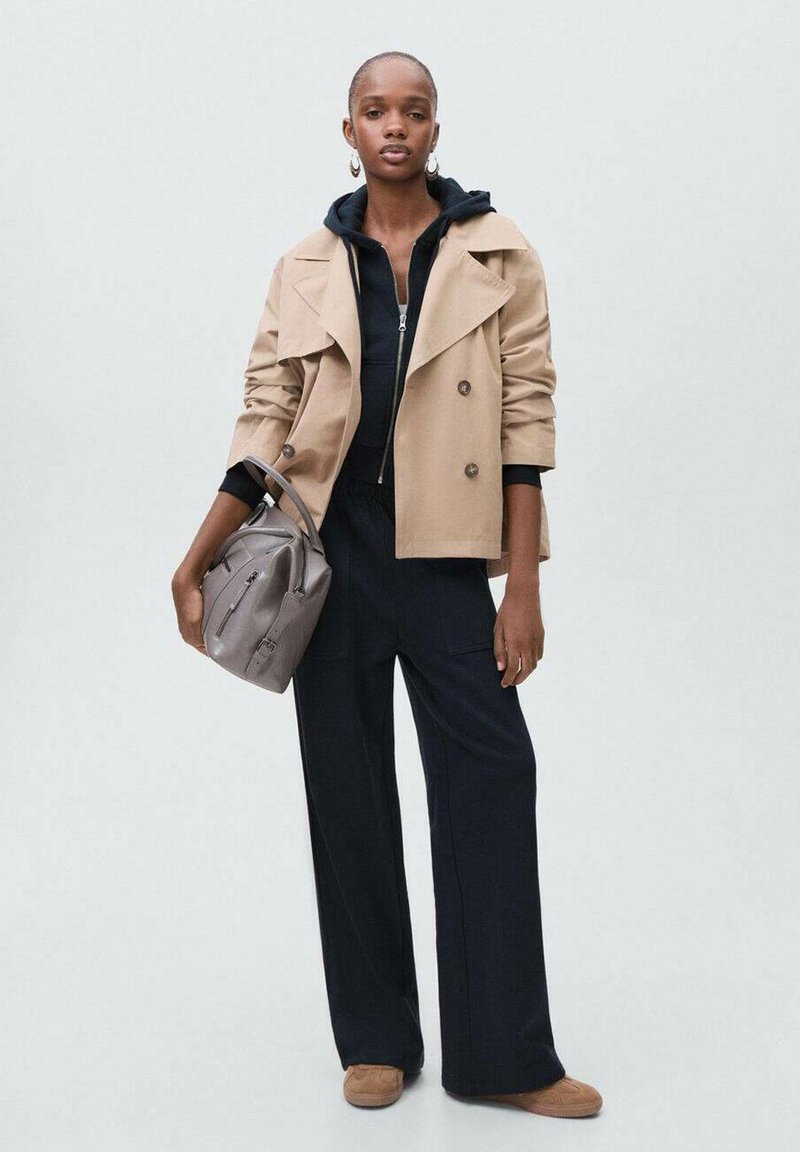 Tan double-breasted trench coat over a black zip hoodie, paired with dark wide-leg pants and light brown slip-on shoes. Gray shoulder bag.