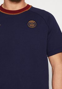 Camiseta de manga corta azul marino con cuello redondo burdeos y costuras doradas, con un logo circular del Paris Saint-Germain en el pecho.