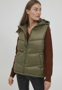 Gilet rembourré vert olive avec fermeture éclair sur le devant, capuche et poches latérales. Il présente un design matelassé, une texture douce et est porté sur un pull en tricot marron.