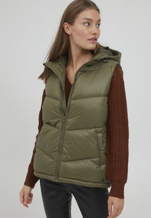 Gilet rembourré vert olive avec fermeture éclair sur le devant, capuche et poches latérales. Il présente un design matelassé, une texture douce et est porté sur un pull en tricot marron.