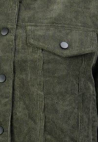 Grønn kordfløyelsjakke med frontdetaljer, vertikale rygger, svarte trykknapper og en klaffelomme på brystet.