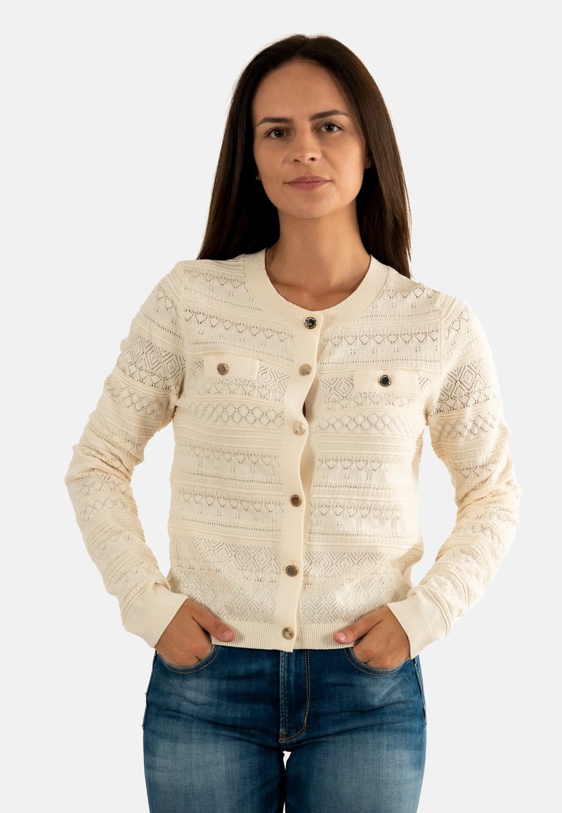 Donna con lunghi capelli castani indossa un cardigan in maglia crema con bottoni e jeans blu, in piedi con le mani nelle tasche su uno sfondo semplice.