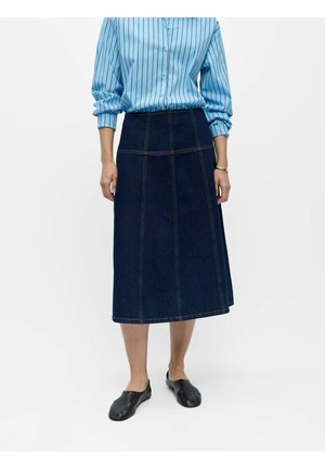 Frau trägt ein blau gestreiftes, geknöpftes Hemd, das in einen dunkelblauen knielangen Jeansrock gesteckt ist, mit schwarzen Slippern.