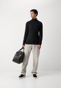 Schwarzer gerippter Rollkragenpullover, hellgraue oversized Hose, schwarze Ledertasche, schwarze Sneaker mit cremefarbenen Sohlen, steht vor einem neutralen Hintergrund.