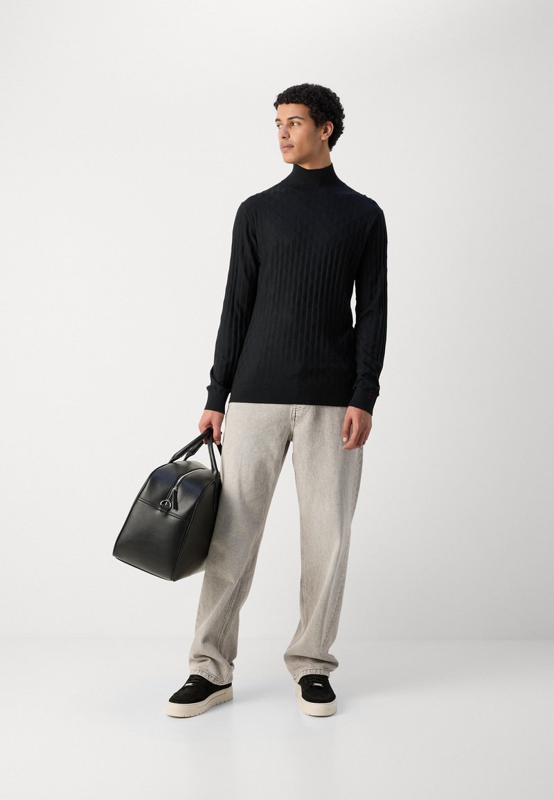 Zwarte geribbelde coltrui, lichtgrijze oversized broeken, zwarte leren tas, zwarte sneakers met crèmekleurige zolen, staand tegen een neutrale achtergrond.