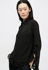 Blusa preta em chiffon com fecho de botão, mangas longas com detalhes em babados e tecido transparentemente, e um corte ajustado.
