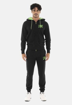Homme portant un sweat à capuche noir zippé et un pantalon assorti avec un logo vert, debout, les mains relâchées le long du corps sur fond blanc.
