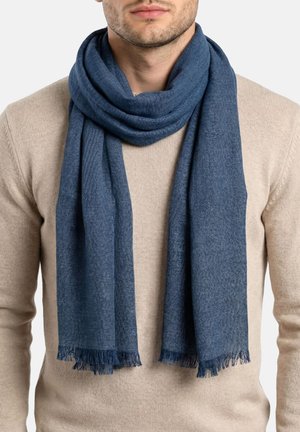 Hombre que lleva un suéter beige y una bufanda tejida azul oscuro con extremos con flecos, envuelta suelta alrededor del cuello sobre un fondo liso.