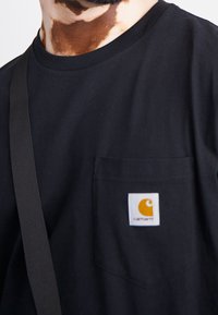 Černé bavlněné tričko s kapsou na hrudi a bílou etiketou s oranžovým logem. Materiál je hladký a má design s kulatým výstřihem.