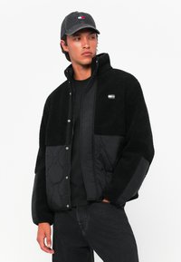 Zwarte jas met fleece en gequiltte panelen, voorzien van een hoge kraag en drukknopen. Zwarte gestructureerde pet met logo-accenten.