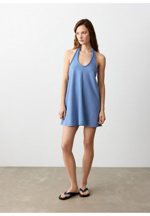 Žena stojaca v modrých šatách bez rukávov s halterneck výstrihom a čiernych žabkách na sivom podklade s jednoduchou bielou stenou v pozadí.