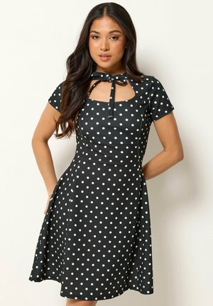 Frau trägt ein schwarzes, knielanges Kleid mit kurzen Ärmeln, weißen Punkten und einer Schleife am Ausschnitt, steht vor einer schlichten Wand.