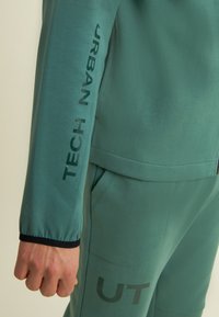 Ensemble athlétique vert composé d'une veste zippée et d'un pantalon. Détails notables : texte imprimé sur les manches et le pantalon, poignets élastiques et tissu texturé.