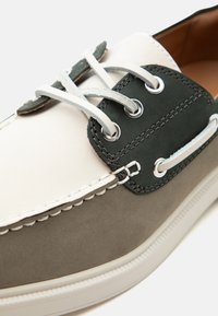 Gros plan d’une chaussure bateau beige, vert foncé et marron avec des lacets blancs et des œillets métalliques sur une semelle blanche.