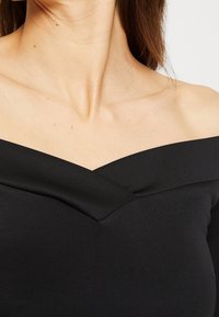 Femme portant un haut noir décolleté sur les épaules, dévoilant le cou et la clavicule, avec de longs cheveux bruns partiellement visibles.