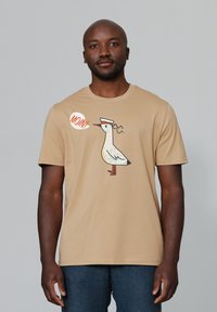 Beige T-shirt met korte mouwen, met een cartoonvogel met een hoed en de tekst "MOIN!" in een spraakbubble. Zachte stof, eenvoudig ontwerp.