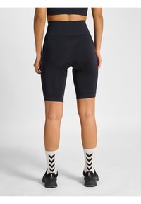 Femme portant un short cycliste noir taille haute, des chaussettes blanches à chevrons noirs et des chaussures de sport noires, debout de dos sur un fond blanc.