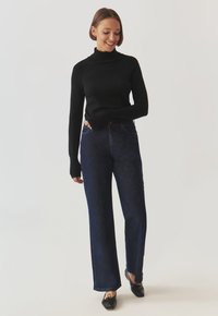 Maglione nero a collo alto con maniche aderenti, abbinato a jeans dritti in denim scuro a vita alta. Il modello indossa scarpe nere con un dettaglio a fiocco.