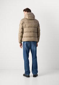 Doudoune beige avec capuche, présentant des sections matelassées. Portée sur un jeans en denim bleu et des chaussures noires. Design simple sans logos visibles.