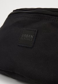 Sac en tissu noir avec une surface texturée, fermeture éclair et un logo carré en relief portant la mention "URBAN CLASSICS" dans le coin.