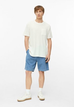 Jeune homme portant un t-shirt blanc à manches courtes, un short en jean bleu, des chaussettes blanches et des baskets blanches, debout devant un fond blanc.