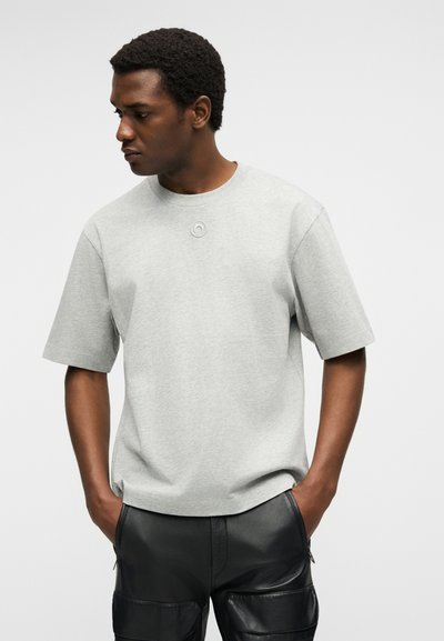 Άνδρας με κοντά σγουρά μαλλιά που φοράει ανοιχτό γκρι κοντομάνικο πουκάμισο και μαύρο δερμάτινο παντελόνι, με τα χέρια στις τσέπες, κοιτάζει προς τα αριστερά του.