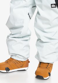Pantalones de nieve azul claro con puños elásticos, combinados con botas de nieve marrones y negras que presentan un diseño texturizado y acentos blancos.