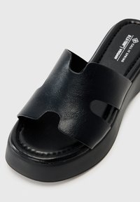 Sandalia vegana negra tipo slide con un diseño de tira curvada y aberturas, suela elevada y textura suave. Construcción minimalista y moderna.
