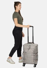 Grau melierter Stoffkoffer mit Rollen, verstellbarem Griff, Seitenriemen und Netzfächern. Der Benutzer trägt schwarze Jeans und helle Turnschuhe und steht seitlich.