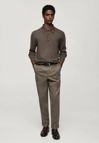 Polo-shirt marron avec un col, trois boutons et des manches longues, associé à un pantalon beige clair et des bottes noires. Design minimaliste.