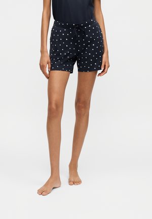 Person trägt marineblaue Shorts mit weißem Herzmuster und Kordelzug in der Taille, steht barfuß vor einem schlichten Hintergrund.