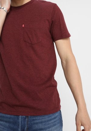 T-shirt bordeaux à manches courtes en tissu doux et texturé avec une poche poitrine et un détail logo subtil. Porté avec un jean en denim bleu.