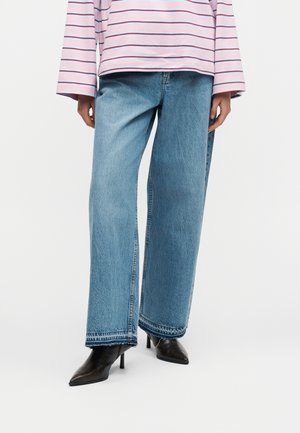 Άτομο που φοράει ανοιχτό γαλάζιο τζιν με φαρδύ πόδι, μαύρες μυτερές μπότες με ψηλό τακούνι και ροζ-λευκό ριγέ πουκάμισο με μακριά μανίκια.