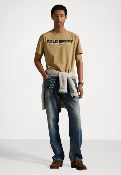 Camiseta de algodón marrón con gráfico de "POLO SPORT", combinada con jeans azul desgastados. Sudadera gris atada a la cintura, zapatos marrones con cordones.
