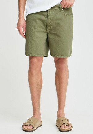 Homme portant un short kaki au genou et des sandales beige à bouts ouverts, debout devant un fond blanc uni.