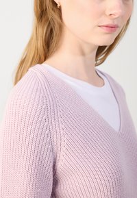 Pulover tricotat de culoare roz deschis, cu decolteu în V și model texturat; purtat peste un tricou alb; are o croială lejeră și mâneci scurte.