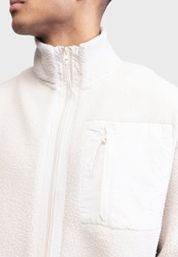 Fleecejacke in Off-White mit hohem Kragen, vollem Frontreißverschluss und einer Reißverschlusstasche auf der Brust. Verfügt über strukturierten Stoff und glatte Akzente.
