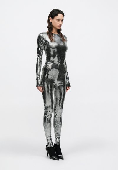 Spīdīgs, pieguļošs ķermeņa tērps no metāliskas auduma, ar garām piedurknēm un apaļu izgriezumu, kombinēts ar melniem potīšu zābakiem uz papēžiem.